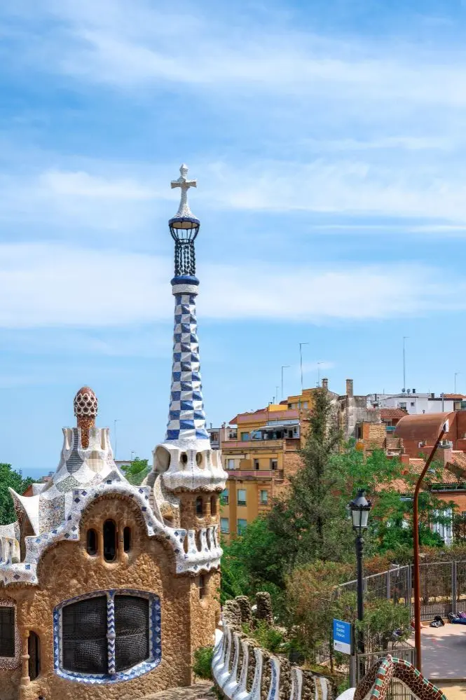 Een uitzicht op een toren in Park Güell, Barcelona, ​​versierd met blauw-witte mozaïeken en bekroond met een kruis, met typische woongebouwen op de achtergrond onder een heldere hemel, ter illustratie van de bezorgdiensten van Landmark Global in Spanje.
